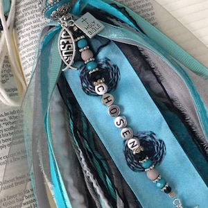 May include: A blue and white fabric bookmark with a silver charm that says "JESUS" and a silver charm that says "HOLY BIBLE". The bookmark also has a silver charm with the word "CHOSEN" spelled out in individual beads. The bookmark is decorated with beads and ribbons.