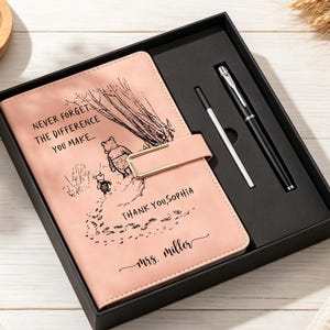 Op de afbeelding: Een lichtroze notitieboek met een goudkleurige metalen sluiting, voorzien van een zwarte lijntekening van Winnie de Poeh en Knorretje, de tekst "NEVER FORGET THE DIFFERENCE YOU MAKE...", "THANK YOU, SOPHIA" en "mrs. miller", inclusief een zwarte pen.