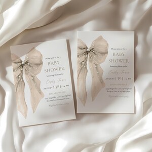 May include: Two baby shower invitations featuring a watercolour bow design. The invitations are white with a light beige bow and pearl accents. Text includes "Baby Shower", the honoree's name, date, time, and location.
