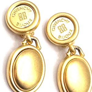 Puede incluir: Un par de pendientes de gota de tono dorado con un diseño circular. La parte superior de cada pendiente presenta un disco circular con el texto "Givenchy Bijoux" en letras doradas. La parte inferior de cada pendiente presenta un disco liso, redondo y de tono dorado.