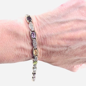 May include: A multi-gemstone link bracelet featuring oval-cut stones in shades of purple, yellow, and green, alternating with small silver-toned links encrusted with tiny clear accent stones. The bracelet has a polished metal finish throughout.
