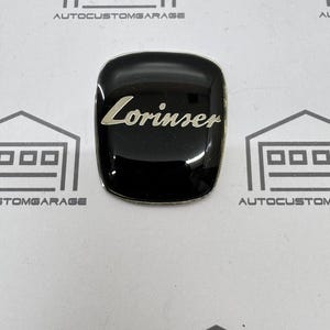 May include: A black, glossy emblem with the word "Lorinser" in silver script. The emblem is square with rounded corners and a silver border. The background features the text "AUTOCUSTOMGARAGE".