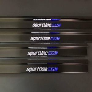 Könnte beinhalten: Ein Set aus vier rechteckigen Türschwellen aus Edelstahl mit polierter Oberfläche. Jede Platte zeigt den Schriftzug sportline in weißen Buchstaben, gefolgt von vier blau beleuchteten Quadraten. Dünne schwarze elektrische Kabel sind angebracht.