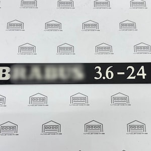 May include: A rectangular black metal emblem featuring the white text BRABUS and the numbers 3.6-24 in a bold, sans-serif font. The surface has a smooth, matte finish, and the lettering is clearly legible against the dark background.