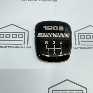 May include: A black gear shift knob with the text "190E evolution" in white. The gear pattern is visible with R, 1, 2, 3, 4, and 5. The knob has a glossy finish and is likely an automotive part.