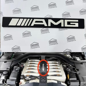 May include: Black AMG emblem with white lettering and stripes. The emblem is rectangular and features the AMG logo. The image also shows the emblem installed on a car engine.