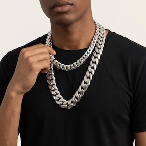 May include: A silver-colored chain necklace with large, interlocking links. The necklace is worn over a black t-shirt. The chain is held by a person's hand.