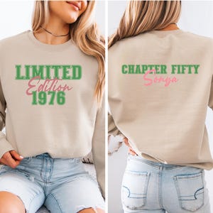 May include: A beige sweatshirt with green and pink text. The front reads "LIMITED Edition 1976". The back of the sweatshirt says "CHAPTER FIFTY Sonya". The sweatshirt is a crew neck style.