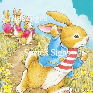 May include: A cartoon illustration of three brown rabbits with pink ears. The rabbits are wearing red and blue jackets and are walking on a path with yellow flowers. The text "Suzie's Signs" is in the top left corner of the image.