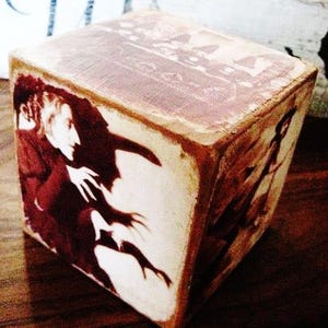 May include: A wooden cube with a vintage image of a witch with a long nose and a shadow behind her. The image is in shades of red, white, and black.