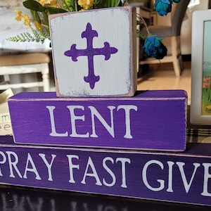 May include: Three wooden blocks stacked on top of each other. The top block is white with a purple cross. The middle block is purple with the word "LENT" in white letters. The bottom block is purple with the words "PRAY FAST GIVE" in white letters.