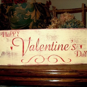 May include: A white wooden sign with red painted lettering that reads "Happy Valentine's Day". The sign has a distressed look and is decorated with red hearts.