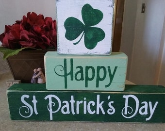 May include: Three wooden blocks stacked on top of each other. The top block is white with a green shamrock painted on it. The middle block is light green with the word "Happy" painted in green. The bottom block is dark green with the words "St Patrick's Day" painted in white.