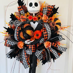 May include: A decorative wreath featuring a white skull head with large black eyes, a black and white pinstriped suit collar, and skeletal hands holding a glittery black and orange pumpkin. It includes orange roses, black and white chequered ribbons, and spider web accents.