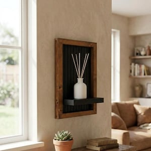 Op de afbeelding: Een houten wandplank met een zwart interieur en een kleine plank. Een witte vaas met riet staat op de plank. Een kleine potplant en boeken staan op een houten tafel eronder.