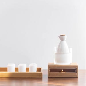 May include: A white ceramic sake bottle with a cork stopper sits on a wooden stand with a small candle burning underneath. The stand is on a wooden table with a tray of six small white ceramic cups.
