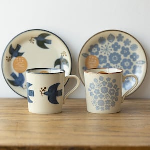 Op de afbeelding: Twee keramische mokken met blauwe en witte bloemen- en vogelontwerpen, elk met een bijpassende schotel. De mokken hebben een crèmekleurige achtergrond.