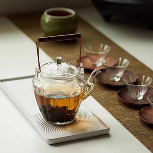 Può includere: Una teiera in vetro trasparente con manico in legno marrone e struttura in metallo, piena di tè. La teiera è appoggiata su un vassoio bianco con un design circolare. Piccole tazzine di vetro e piattini di legno sono disposte nelle vicinanze.