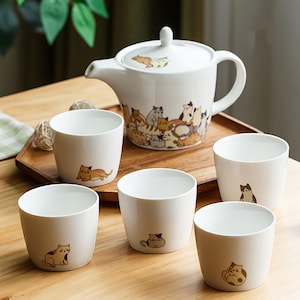 Peut inclure: Une théière en céramique blanche et cinq tasses à thé avec un motif de chat. La théière a un couvercle et une poignée. Les tasses à thé ont un motif de chat sur le devant. La théière et les tasses à thé sont sur un plateau en bois.