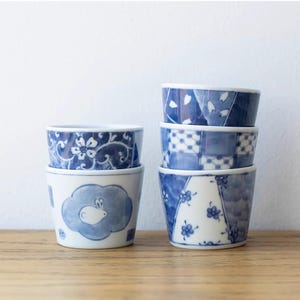 Peut inclure: Un ensemble de quatre tasses à thé en céramique bleu et blanc avec des motifs différents. Les tasses sont empilées les unes sur les autres. Les motifs comprennent un motif floral, un motif à carreaux et un motif avec un lapin dans un nuage.