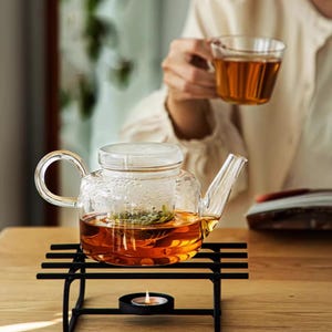 以下が含まれることがあります： カーブしたハンドルとガラスの蓋が付いた透明なガラスのティーポットで、お茶が入っています。ティーポットは、下に小さなろうそくが灯された黒い金属製のスタンドの上にあります。背景には、お茶のグラスを持っている人がいます。