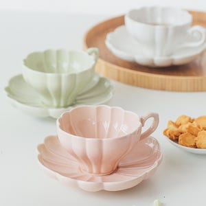 Peut inclure: Ensemble de tasses et soucoupes aux couleurs pastel. Les tasses et les soucoupes ont un bord festonné. Les couleurs comprennent le rose, le vert et le blanc. Les tasses sont sur une surface blanche, avec un plateau en bois en arrière-plan.