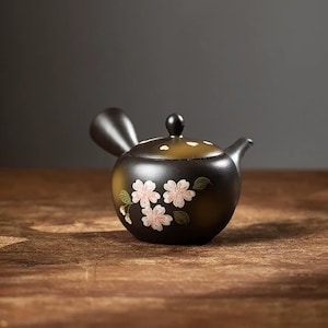 Könnte beinhalten: Kleine, schwarze Keramik-Teekanne mit rundem Körper und gebogenem Ausguss. Die Teekanne hat einen Deckel mit einem kleinen Knauf und ist mit rosa Kirschblüten und grünen Blättern verziert. Sie steht auf einer Holzoberfläche.