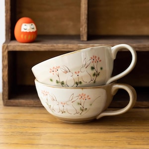 May include: Two white ceramic mugs with handles, stacked on top of each other. Both mugs feature a design of pink flowers and white rabbits. The mugs are on a wooden surface.