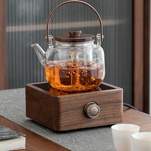 Może przedstawiać: Szklany czajnik z drewnianym pokrywką i uchwytem stoi na drewnianym elektrycznym podgrzewaczu do herbaty. Czajnik jest wypełniony brązową herbatą, a podgrzewacz ma pokrętło z przodu.