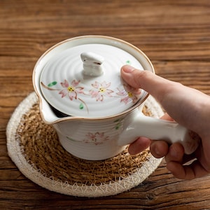 May include: White ceramic teapot with a floral design and a small rabbit on the lid. The teapot is sitting on a woven mat.