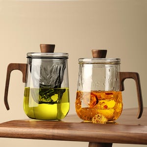 May include: Two glass tea mugs with wooden handles and lids. One mug contains green tea, the other contains a golden-colored tea. Both mugs have a clear glass design with a stainless steel tea infuser. The mugs are sitting on a wooden surface.