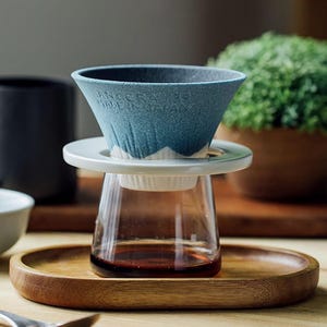 Op de afbeelding: Een blauwe keramische pour-over koffiemaker met een witte rand en een heldere glazen karaf. De karaf staat op een houten dienblad. De koffiemaker heeft de tekst "ANGER MODERN" aan de zijkant.
