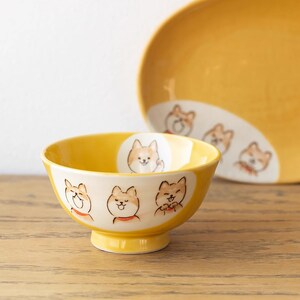 Peut inclure: Un bol en céramique jaune avec un intérieur blanc présente quatre chiens Shiba Inu de dessin animé avec des expressions différentes. Le bol a une forme arrondie et un petit pied.