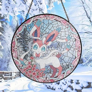 Puede incluir: Vidriera redonda con un diseño de personaje Sylveon. La obra de arte presenta una paleta de colores blanco, rosa y azul, con un borde floral y una cadena para colgar. El fondo es una escena invernal.
