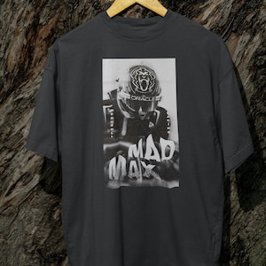May include: A dark grey t-shirt featuring a black and white image of a racing driver wearing a helmet with the word "ORACLE" on it. The text "MAD MAX" is printed below the image.