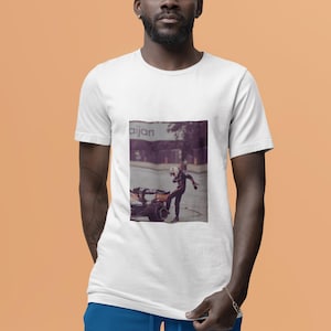 May include: A white t-shirt with a graphic of a person in a racing suit standing next to a race car. The text "aijan" is visible on the graphic.
