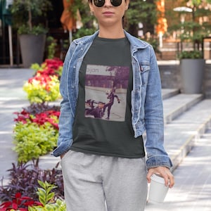 May include: A dark gray t-shirt with a graphic print of a person riding a small motorcycle. The graphic is in a vintage style and includes the word "aijan".