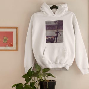 K&ouml;nnte beinhalten: Wei&szlig;er Hoodie mit einem Grafikdruck einer Person in schwarzer Kleidung, die vor einem Geb&auml;ude mit dem Wort "Bijan" steht.