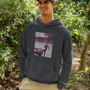 K&ouml;nnte beinhalten: Ein dunkelgrauer Kapuzenpullover mit einem Grafikdruck einer Person in einem Rennanzug, die einen Reifen tritt. Der Text "aijan" ist auf dem Grafikdruck gedruckt.