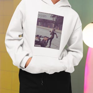 K&ouml;nnte beinhalten: Wei&szlig;er Hoodie mit einem schwarzen und orangefarbenen Formel-1-Rennwagen und einem Fahrer, der auf der Vorderseite aufgedruckt ist. Der Text "aijan" ist auf dem Geb&auml;ude im Hintergrund sichtbar.