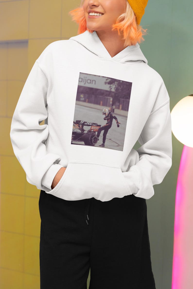 K&ouml;nnte beinhalten: Wei&szlig;er Hoodie mit einem schwarzen und orangefarbenen Formel-1-Rennwagen und einem Fahrer, der auf der Vorderseite aufgedruckt ist. Der Text "aijan" ist auf dem Geb&auml;ude im Hintergrund sichtbar.