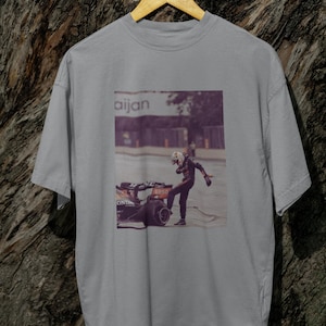 May include: A gray t-shirt with a graphic print of a race car driver standing next to a race car. The text "aijan" is printed on the shirt.