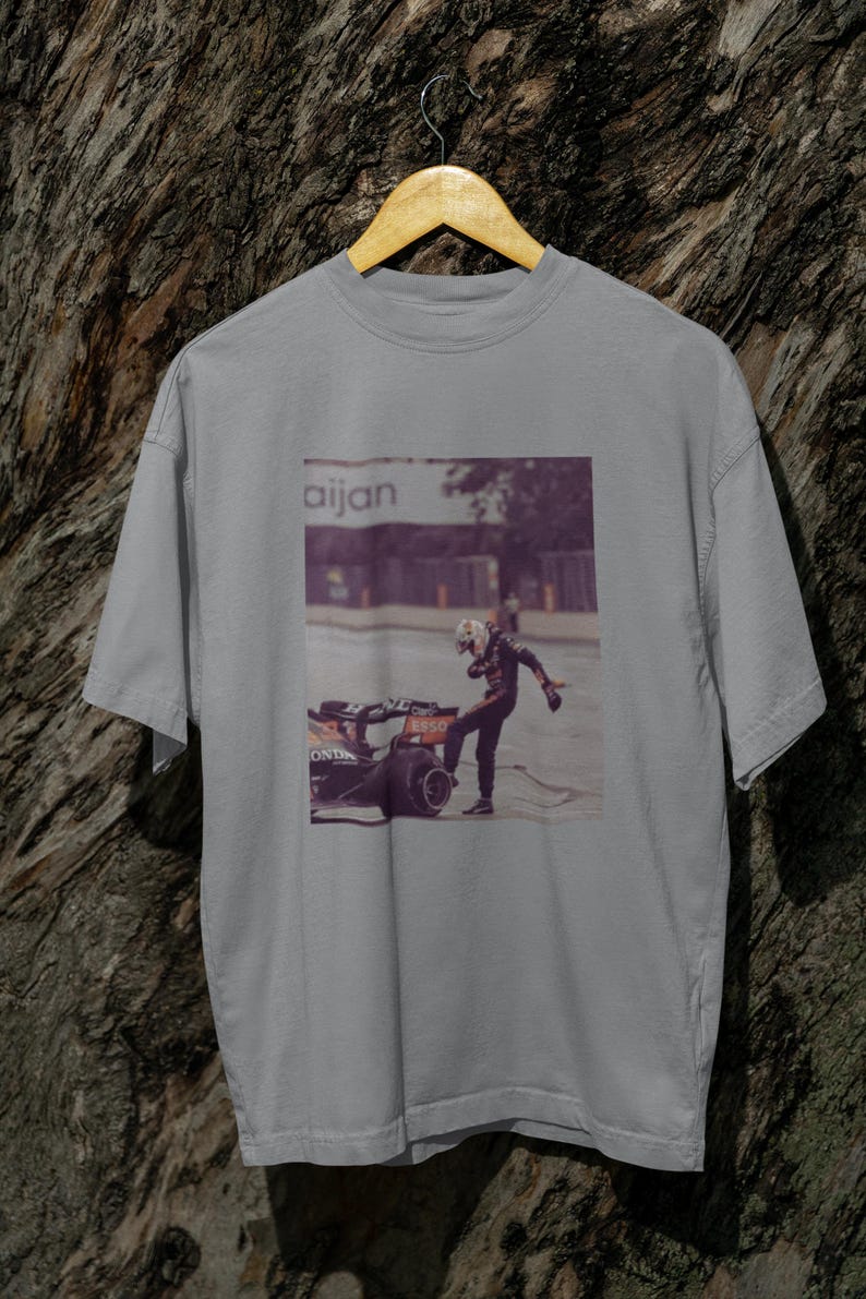 May include: A gray t-shirt with a graphic print of a race car driver standing next to a race car. The text "aijan" is printed on the shirt.