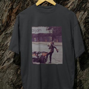 May include: A dark gray t-shirt with a graphic print of a race car driver in a helmet and racing suit standing next to a race car. The text "aijan" is printed on the graphic.