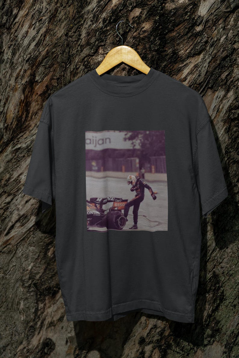 May include: A dark gray t-shirt with a graphic print of a race car driver in a helmet and racing suit standing next to a race car. The text "aijan" is printed on the graphic.
