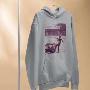 K&ouml;nnte beinhalten: Ein grauer Kapuzenpullover mit einem Grafikdruck eines Rennfahrers, der neben einem Rennwagen steht. Der Text "aijan" ist auf dem Grafikdruck gedruckt.