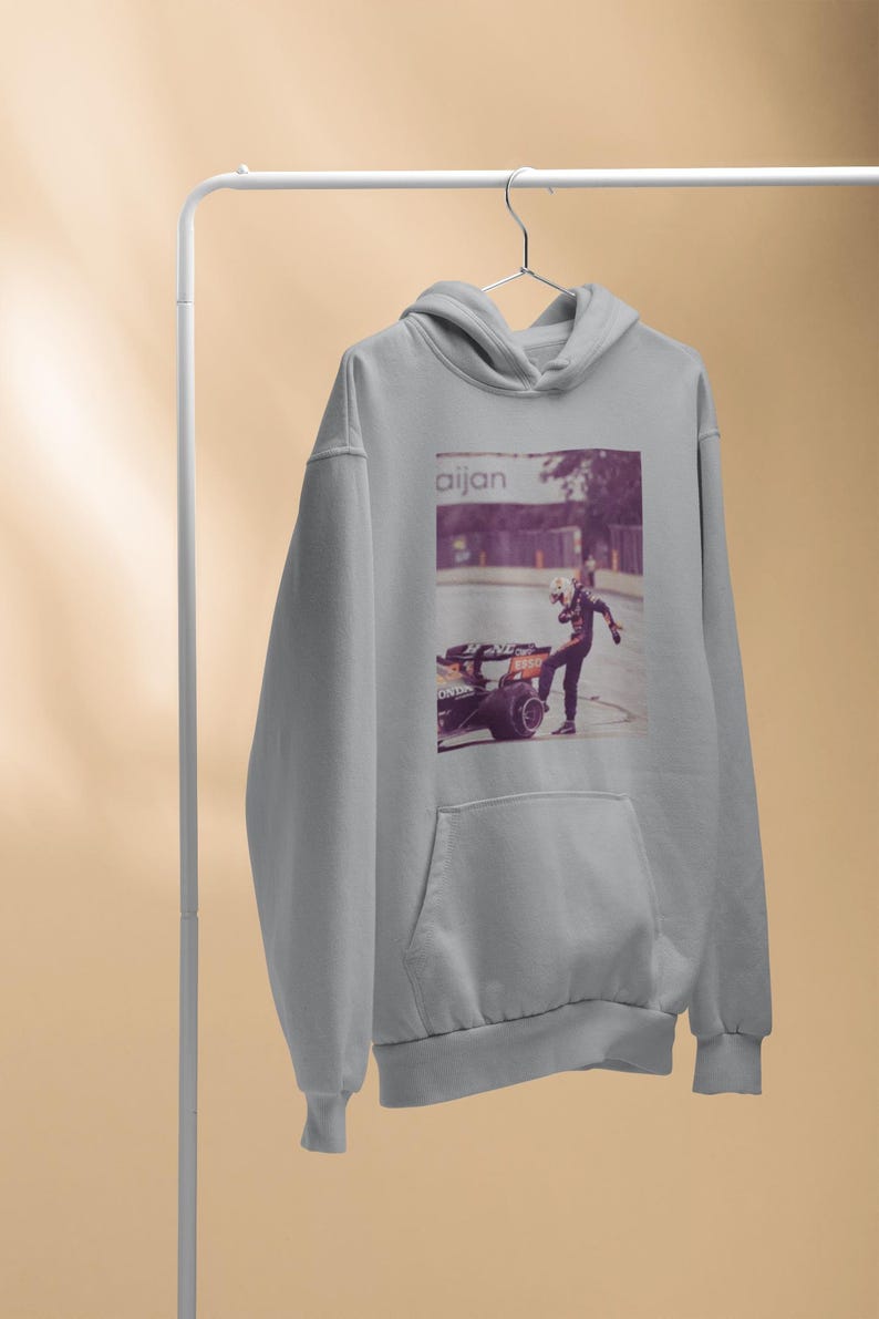 K&ouml;nnte beinhalten: Ein grauer Kapuzenpullover mit einem Grafikdruck eines Rennfahrers, der neben einem Rennwagen steht. Der Text "aijan" ist auf dem Grafikdruck gedruckt.