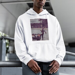 K&ouml;nnte beinhalten: Ein wei&szlig;er Hoodie mit einem Grafikdruck einer Person in einem Rennanzug, die neben einem Rennwagen steht. Die Grafik ist in Violett- und Schwarzt&ouml;nen gehalten. Der Hoodie hat eine Kapuze mit Kordelzug und eine K&auml;ngurutasche.