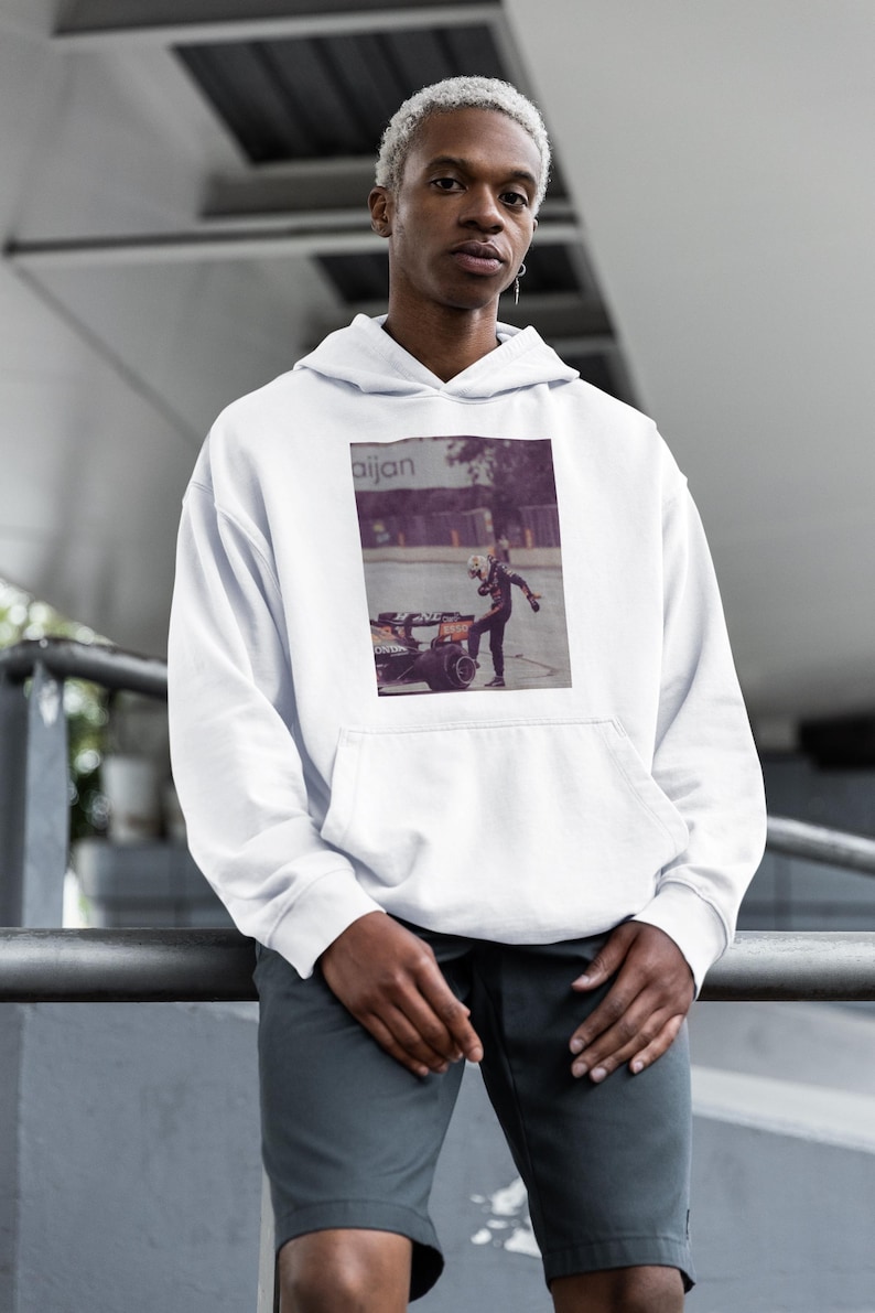 K&ouml;nnte beinhalten: Ein wei&szlig;er Hoodie mit einem Grafikdruck einer Person in einem Rennanzug, die neben einem Rennwagen steht. Die Grafik ist in Violett- und Schwarzt&ouml;nen gehalten. Der Hoodie hat eine Kapuze mit Kordelzug und eine K&auml;ngurutasche.