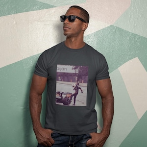 May include: A man wearing a dark gray t-shirt with a vintage photo of a person in a racing suit and a car. The photo has the word "Bijan" printed on it.